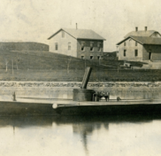 LIGHTNING (HMCo. #20), the United States Navy’s first torpedo boat in Newport waters, 1876. Sepia tone photograph. Workshop buildings in the background. Torpedo boat in the foreground. Portside View of boat.