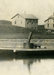 LIGHTNING (HMCo. #20), the United States Navy’s first torpedo boat in Newport waters, 1876. Sepia tone photograph. Workshop buildings in the background. Torpedo boat in the foreground. Portside View of boat.