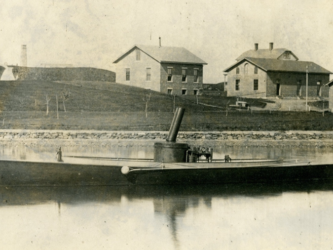 LIGHTNING (HMCo. #20), the United States Navy’s first torpedo boat in Newport waters, 1876. Sepia tone photograph. Workshop buildings in the background. Torpedo boat in the foreground. Portside View of boat.