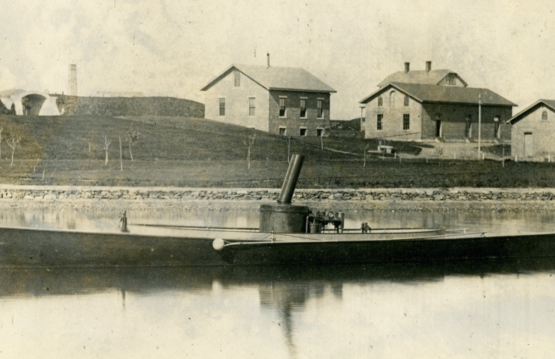LIGHTNING (HMCo. #20), the United States Navy’s first torpedo boat in Newport waters, 1876. Sepia tone photograph. Workshop buildings in the background. Torpedo boat in the foreground. Portside View of boat.
