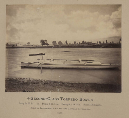 Sepia photo of torpedo boat moored near the shoreline. Land is visible in the background on the horizon. Text at bottom of photo reads: Second Class Torpedo Boat. Length, 67ft 0in. Beam, 8ft 6in. Draught, 4ft 3in. Speed 18.2 knots. Built by Thornycroft & Co. for the Austrian Government. 1875