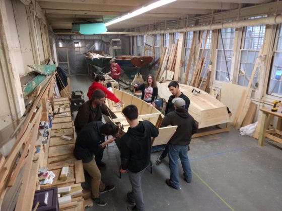 Highschool students surround a ten foot wooden skiff they are building